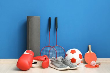 Spor ayakkabılar, yoga minderi, masa tenisi raketleri, boks eldivenleri, badminton raketleri ve mavi duvarın yanındaki yerde mekikçikler.