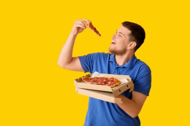 Elinde karton kutu ve sarı arka planda lezzetli pepperonili pizza tutan genç adam.