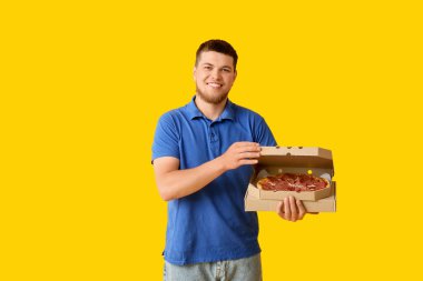 Sarı arka planda lezzetli pepperonili pizzayla dolu karton kutu tutan mutlu genç adam.
