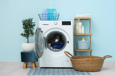 Mavi duvarın yanında rafı, sepeti ve deterjanı olan modern çamaşır makinesi.