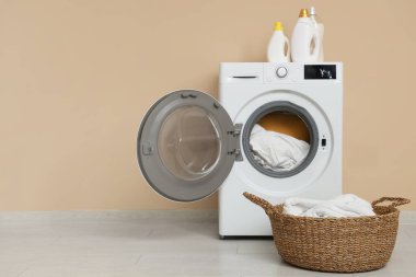 Bej duvarın yanında çamaşır, sepet ve deterjan olan modern çamaşır makinesi.