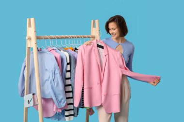 Farklı giysileri olan, mavi arka planda ceketi seçen güzel genç bir kadın.