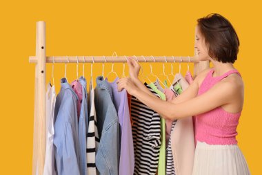 Sarı arka planda farklı giysileri olan, rafların yanında güzel genç bir kadın.