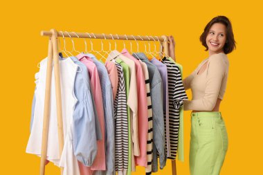 Sarı arka planda farklı giysileri olan, rafların yanında güzel genç bir kadın.