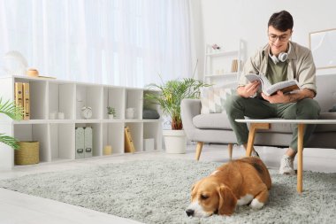 Kulaklıklı genç adam, evdeki sevimli Beagle köpeğinin yanındaki kanepede dergi okuyor.