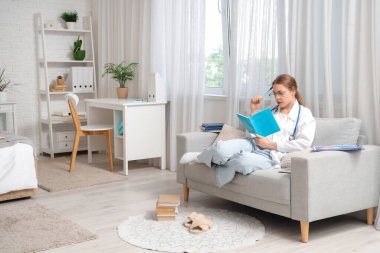 Bayan tıp öğrencisi evdeki kanepede defterle ders çalışıyor.