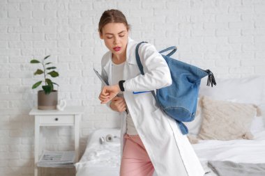 Laptoplu ve sırt çantalı bir tıp öğrencisi evde kol saatine bakıyor.