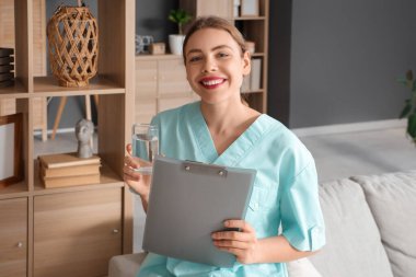 Genç bir tıp öğrencisi, elinde dosya ve bir bardak suyla evdeki kanepede oturuyor.