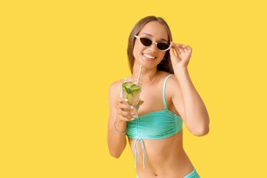 Sarı arka planda bir bardak mojito ile mayo giymiş güzel, genç bir kadın.