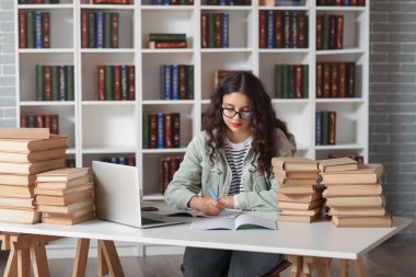 Gözlüklü kız öğrenci dizüstü bilgisayarı ve kütüphanedeki kitap yığınıyla masadaki not defterine yazıyor.