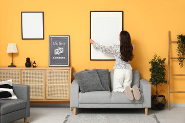 Evdeki turuncu duvara boş bir çerçeve asan genç bir kadın. Arka plan