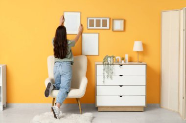 Oturma odasındaki koltuğun yanındaki turuncu duvara boş bir çerçeve asan genç kadın. Arka plan