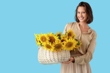 Hasır sepeti ve mavi arka planda bir buket ayçiçeğiyle güzel, genç ve mutlu bir kadın.