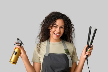 Gri arka planda saç düzleştiricisi ve spreyiyle güzel, mutlu, Afro-Amerikan bir kuaför.