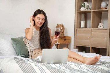 Şarap ve dizüstü bilgisayarlı genç Asyalı kadın yatak odasında randevusu var.