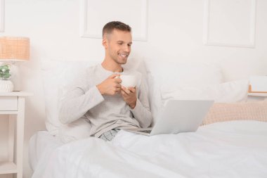 Elinde bir fincan kahve ve dizüstü bilgisayarla yatak odasında randevusu olan genç bir adam.