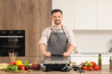 Genç ve mutlu adam mutfaktaki modern elektrikli ızgarada taze sebzeler pişiriyor.