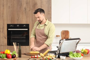 Genç adam mısır koçanı keserek mutfaktaki modern elektrikli ızgarada yemek yapıyor.