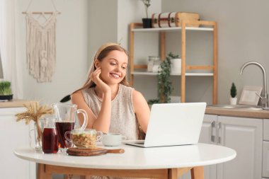 Okroshka 'lı genç bir kadın mutfaktaki masada dizüstü bilgisayar kullanıyor.