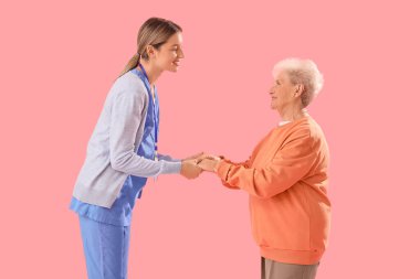 Pembe arka planda hemşireyle el ele tutuşan kıdemli kadın.
