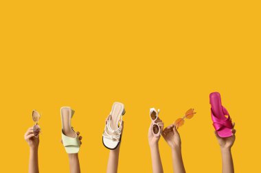 Şık güneş gözlüklü kadın elleri ve sarı arka planda farklı sandaletler.