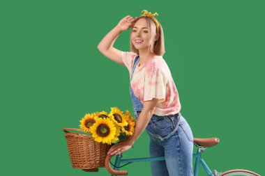 Şık makyajı, bisikleti ve sepetinde ayçiçekleri olan yeşil arka planda güzel, genç ve mutlu bir kadın.
