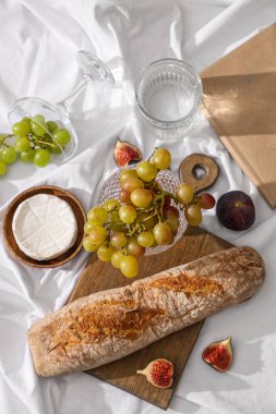 İncirli olgun üzümler, peynirli camembert peyniri, baget ve beyaz kumaş arka planda bir bardak su.