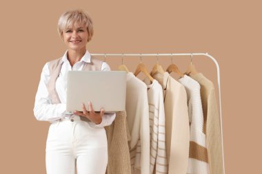 Mutlu olgun kadın stilisti bej arka planda şık elbiseleri olan dizüstü bilgisayar kullanıyor.