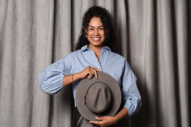 Gri kumaş arka planda şık mücevherleri ve şapkası olan güzel, mutlu, Afro-Amerikan bir kadın.