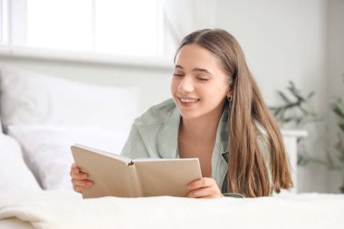 Pijamalı genç ve güzel bir kadın evde kitap okuyor.