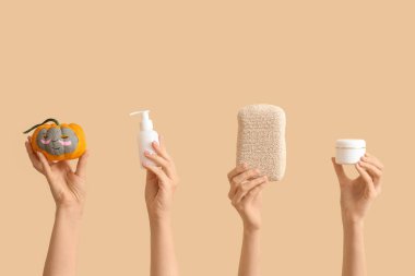 Yüzü çizilmiş balkabağı tutan kadın elleri, kilden maske, banyo süngeri, bej arka planda şişe ve bir kavanoz kozmetik ürünü.