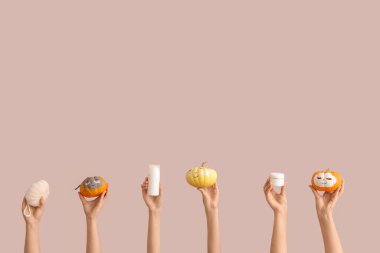 Yüzü çizilmiş balkabaklarını tutan kadın elleri, kilden maskeler, banyo süngeri, şişe ve pembe arka planda kozmetik ürünler kavanozu.