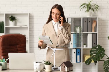 Ofiste cep telefonuyla konuşan kadın mühendis, defter ve rüzgar türbini modeli.