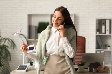 Rüzgâr türbini modeli olan kadın mühendis ofiste cep telefonuyla konuşuyor.