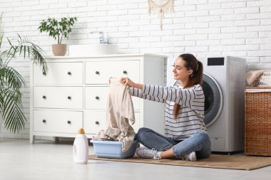 Güzel, genç, mutlu bir kadın. Temiz çamaşır leğeni ve deterjanıyla evde oturuyor.