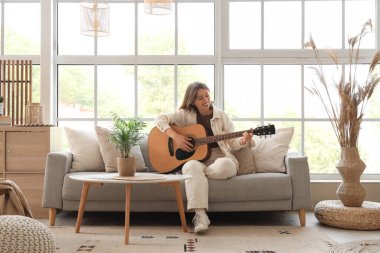 Oturma odasındaki yumuşak koltukta gitar çalan genç bir kadın.