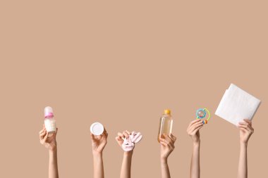 Bej arka planda doğum çantası için bebek eşyaları taşıyan birçok kadın eli var.