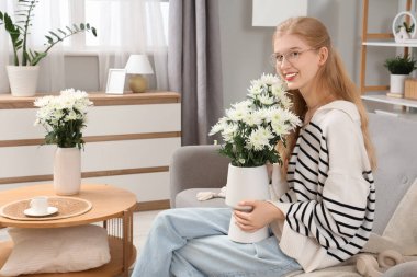 Evdeki kanepede oturan, kulaklıklı, beyaz kasımpatı buketli, genç ve güzel bir kadın.