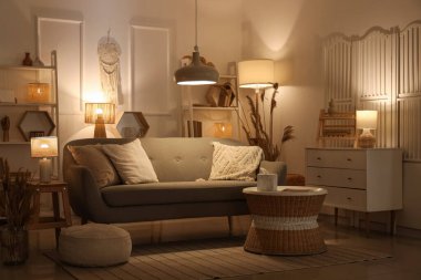 Rahat bir kanepe, çekmeceler, kahve masası, otlar ve oturma odasının içinde parlayan lambalar.