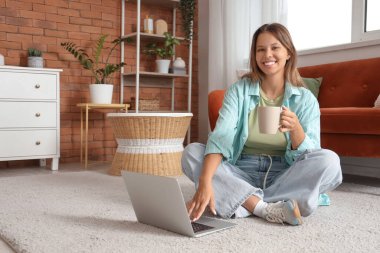 Evde dizüstü bilgisayar kullanan çay içen genç bir kadın.