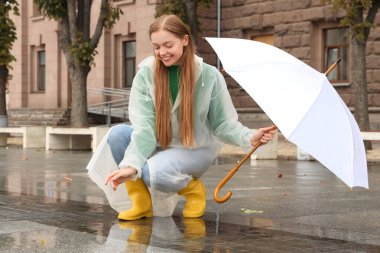 Yağmurluk ve şemsiyeli çizmeli güzel, mutlu bir kadın yağmurlu bir günde dışarıda oturuyor.