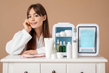 Bej arka planda kozmetik ürünleri için küçük buzdolabı olan güzel genç bir kadın.