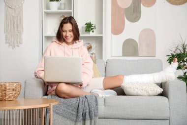Kırık bacaklı genç kadın evdeki kanepede dizüstü bilgisayar kullanıyor.