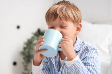 Pijamalı, elinde çay olan, evde yatağında oturan tatlı bir çocuk.