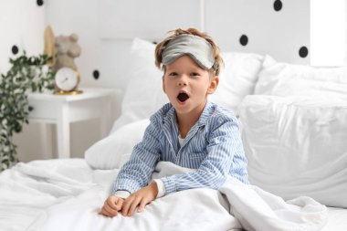 Pijamalı, uyku maskesiyle evdeki yatakta esneyen küçük şirin bir çocuğun sabahı.