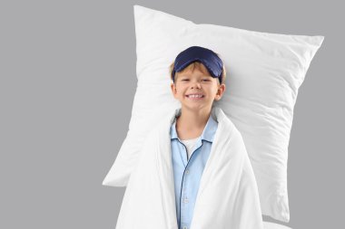 Gri arka planda uyku maskesi, yastığı ve battaniyesi olan pijamalı şirin, mutlu bir çocuk.