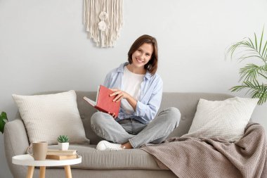 Beyaz duvarın yanındaki kanepede kitap okuyan güzel bir kadın.