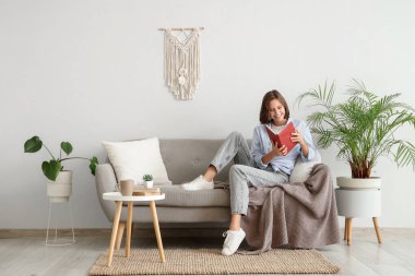 Beyaz duvarın yanındaki kanepede kitap okuyan güzel bir kadın.