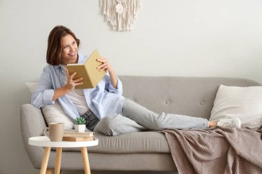 Beyaz duvarın yanındaki kanepede kitap okuyan güzel bir kadın.