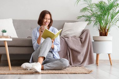 Evdeki kanepenin yanında elinde çay fincanı olan güzel genç bir kadın kitap okuyor.